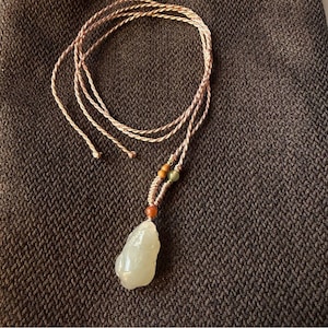 Op de afbeelding: Een lichtgroene jade hangerketting met een gevlochten koord. De hanger heeft een onregelmatige vorm en hangt aan een beige koord. Kleine, kleurrijke kralen sieren het koord bij de hanger. De ketting wordt gepresenteerd op een donkerbruin, getextureerd oppervlak.
