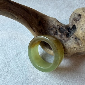 May include: A smooth, wide band ring crafted from translucent stone, featuring a gradient of pale green to warm amber tones. The ring has a polished, rounded surface and a thick, solid construction, showcasing natural color variations throughout.