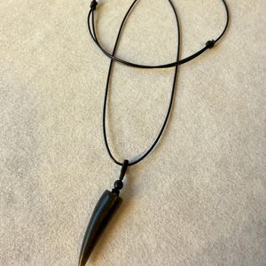 May include: A necklace featuring a smooth, dark, horn-shaped pendant with a subtle amber-colored tip. The pendant is suspended from a thin, black cord that includes a small black bead above the pendant and adjustable sliding knots for length.