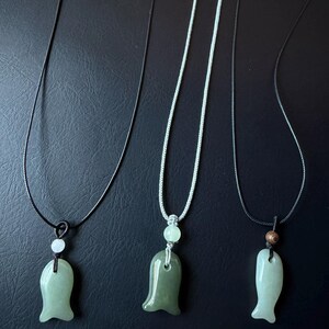 May include: Three jade pendant necklaces. Each necklace features a different shaped jade pendant in shades of green. The pendants are suspended from black and white cords. The background is a dark surface.