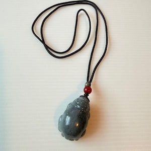 Könnte beinhalten: Grauer Jade-Anhänger in Form eines Frosches, an einer schwarzen Kordelkette. Eine rote Perle akzentuiert die Kordel in der Nähe des Anhängers. Der Frosch-Anhänger hat eine glatte, polierte Oberfläche.