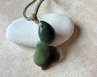 Puede incluir: Un collar con colgante de jade verde oscuro con una forma orgánica única. El colgante está suspendido de un cordón trenzado en tonos dorados, turquesa y marrón. El collar se muestra sobre una piedra blanca sobre un fondo neutro.