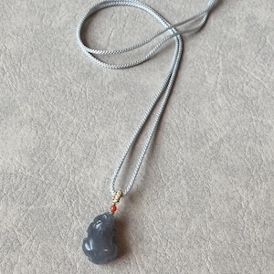 Op de afbeelding: Een grijze jade hangerketting met een gesneden figuur. De hanger hangt aan een lichtgrijs koord met goudkleurige accenten en een kleine rode kraal. De ketting wordt gepresenteerd op een neutraal gekleurd oppervlak.
