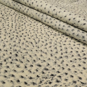 May include: Close-up of a beige ostrich leather fabric with a textured surface. The leather has a pattern of small, dark brown dots.