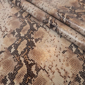 May include: Close-up of a beige and brown snakeskin fabric with a subtle shimmer. The fabric has a textured pattern that resembles snake scales.