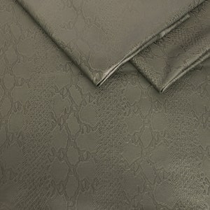 Può includere: Primo piano di un tessuto verde con un motivo a pelle di serpente. Il tessuto è piegato su se stesso, mostrando la trama del materiale.