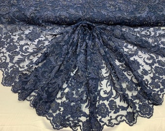 May include: A close-up of a navy blue lace fabric with a floral pattern and beaded accents. The fabric has a scalloped edge.