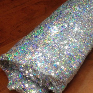 May include: A roll of iridescent silver fabric covered in sequins. The sequins are small and densely packed, creating a shimmering effect.
