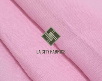 May include: Close-up of a light pink fabric with a subtle texture. The fabric is draped, creating soft folds and shadows. The logo "LA CITY FABRICS" is printed in white below a geometric design.
