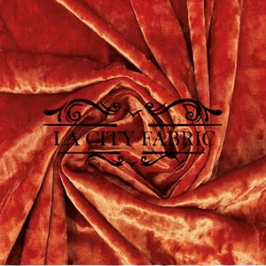 May include: Close-up of a rust-colored velvet fabric with a subtle sheen. The fabric is draped and folded, creating a soft, luxurious look. The text "LA City Fabric" is visible in the center of the image.