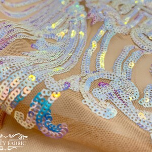 Op de afbeelding: Iridescent witte pailletten op een doorschijnend beige mesh-stof. De pailletten zijn gerangschikt in een draaiende vorm.
