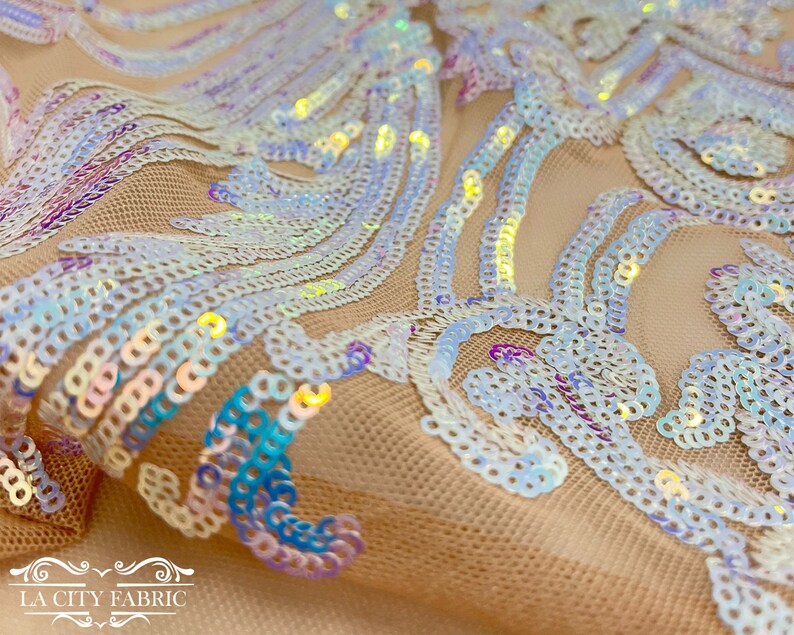 Op de afbeelding: Iridescent witte pailletten op een doorschijnend beige mesh-stof. De pailletten zijn gerangschikt in een draaiende vorm.