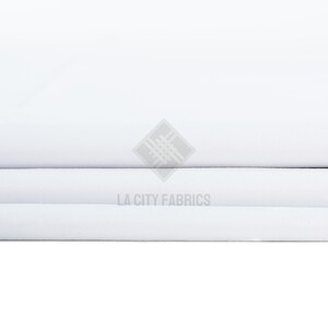 May include: A stack of folded white fabric with a smooth, matte texture. The fabric is layered in horizontal folds. A semi-transparent watermark logo featuring a geometric diamond shape and the text LA CITY FABRICS is centered on the image.