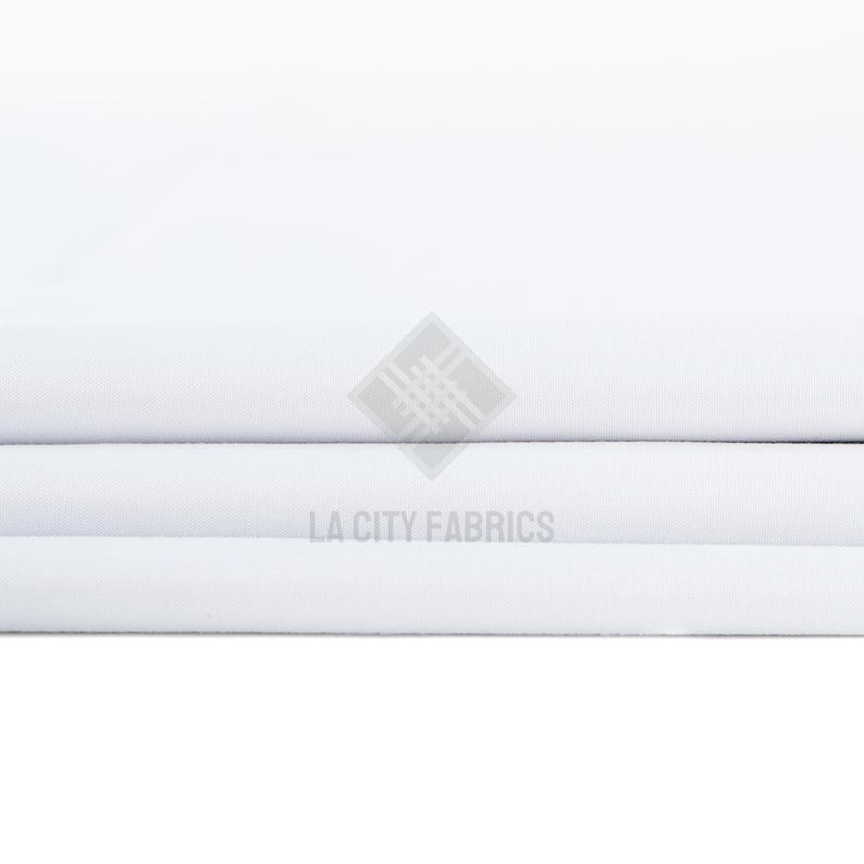 May include: A stack of folded white fabric with a smooth, matte texture. The fabric is layered in horizontal folds. A semi-transparent watermark logo featuring a geometric diamond shape and the text LA CITY FABRICS is centered on the image.