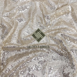 May include: A close-up of a champagne-colored fabric with sequins. The fabric is wrinkled and has a shimmery, iridescent finish.