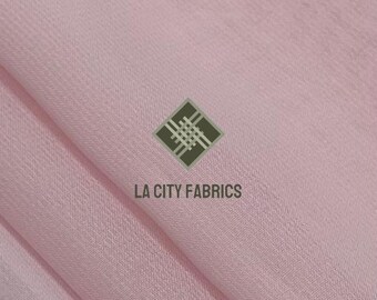 May include: A close-up of a light pink fabric with visible texture and folds. The fabric has a subtle sheen. The logo "LA CITY FABRICS" is printed in white below a green and white geometric design.