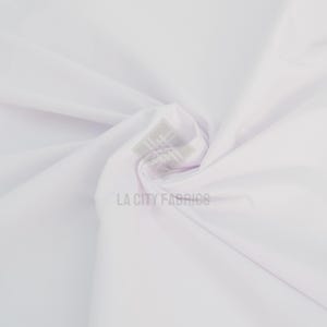 Puede incluir: Una tela blanca sólida con una textura suave, que presenta pliegues y drapeados suaves en todo el material. Un logotipo de marca de agua semitransparente con el texto LA CITY FABRICS está centrado en la superficie de la tela.