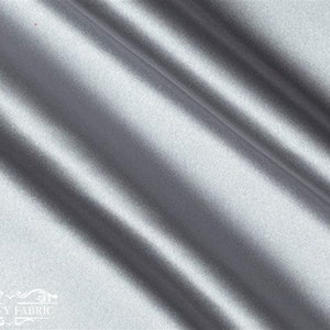 May include: A smooth, silver satin fabric with a subtle sheen. The fabric is folded in a diagonal pattern, showcasing its drape and texture.