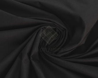 Può includere: Un tessuto nero a tinta unita con una consistenza liscia e opaca, disposto in un motivo a spirale centrale che crea pieghe profonde e ombre morbide sulla superficie. È visibile un logo semitrasparente con la scritta LA CITY FABRICS.