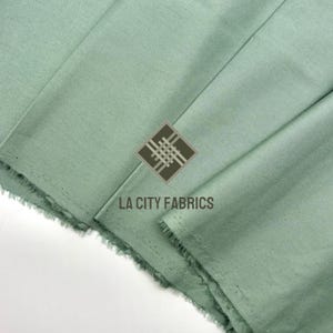 Könnte beinhalten: Nahaufnahme eines gefalteten, hellgrünen Stoffes mit ausgefranstem Rand. Der Stoff hat eine subtile Textur und ist leicht zerknittert. Das Logo "LA CITY FABRICS" ist sichtbar. Der Stoff scheint ein mittelschweres Material zu sein, das sich für Kleidung oder Heimdekoration eignet.