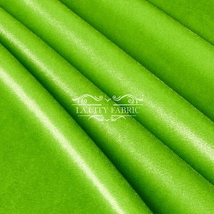 May include: Close-up of a bright green satin fabric. The fabric has a smooth, silky texture and a subtle sheen. The fabric is folded in a way that shows the drape and flow of the material. The text "LA CITY FABRIC" is visible in the center of the image.