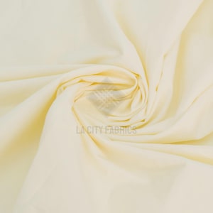 Peut inclure: Un tissu de couleur crème clair uni avec une texture lisse, disposé en un pli en spirale central. Le tissu présente un filigrane en surimpression au centre qui indique LA CITY FABRICS dans une police sans empattement claire sur les plis.