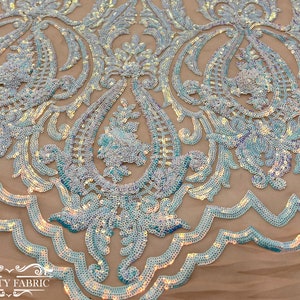 Op de afbeelding: Een close-up van iriserende blauwe pailletten die op een transparante stof zijn genaaid in een paisley-patroon. De pailletten zijn gerangschikt in een bloemmotief met een gegolfde rand.
