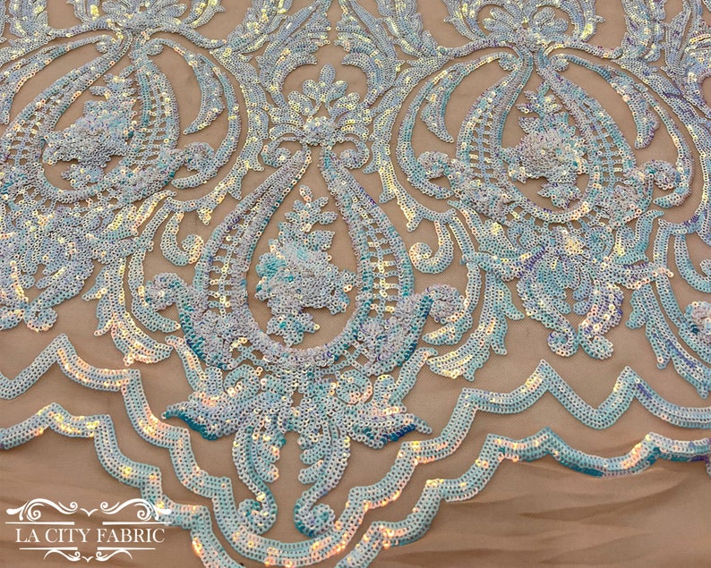 Op de afbeelding: Een close-up van iriserende blauwe pailletten die op een transparante stof zijn genaaid in een paisley-patroon. De pailletten zijn gerangschikt in een bloemmotief met een gegolfde rand.