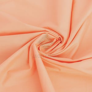Puede incluir: Una tela de color melocotón sólido con una textura suave y mate, dispuesta en un pliegue en espiral central. La tela presenta el texto de marca de agua semitransparente LA CITY FABRICS impreso en el centro del patrón en espiral.