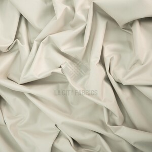May include: A solid off-white fabric with a smooth, matte texture, draped in deep folds and creases. A semi-transparent watermark logo featuring a grid pattern and the text LA CITY FABRICS is centered on the material surface.