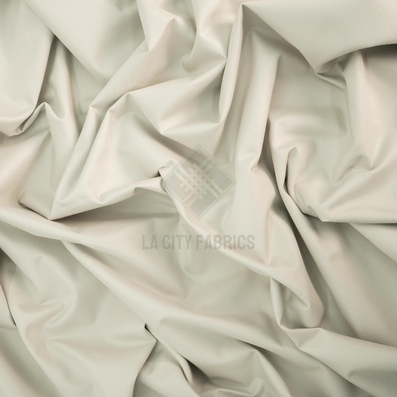 May include: A solid off-white fabric with a smooth, matte texture, draped in deep folds and creases. A semi-transparent watermark logo featuring a grid pattern and the text LA CITY FABRICS is centered on the material surface.