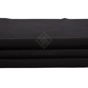 May include: A solid black fabric folded into horizontal layers, featuring a smooth, matte texture. A watermark logo with the text LA CITY FABRICS is centered on the fabric, displaying a diamond-shaped icon composed of intersecting lines.