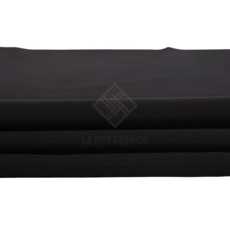 May include: A solid black fabric folded into horizontal layers, featuring a smooth, matte texture. A watermark logo with the text LA CITY FABRICS is centered on the fabric, displaying a diamond-shaped icon composed of intersecting lines.