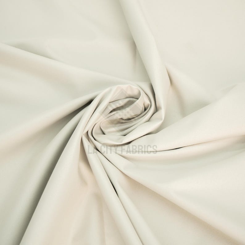 May include: A solid off-white fabric with a smooth, matte texture, arranged in a central spiral fold that creates deep, flowing shadows and highlights across the surface. The text LA CITY FABRICS is visible in the center of the fabric.