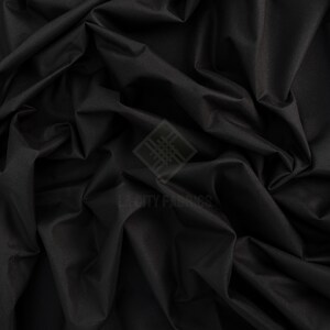 May include: A solid black fabric with a smooth, matte texture and deep, flowing folds. A semi-transparent watermark logo featuring a geometric diamond shape and the text CITY FABRICS is centered on the surface of the dark, draped material.