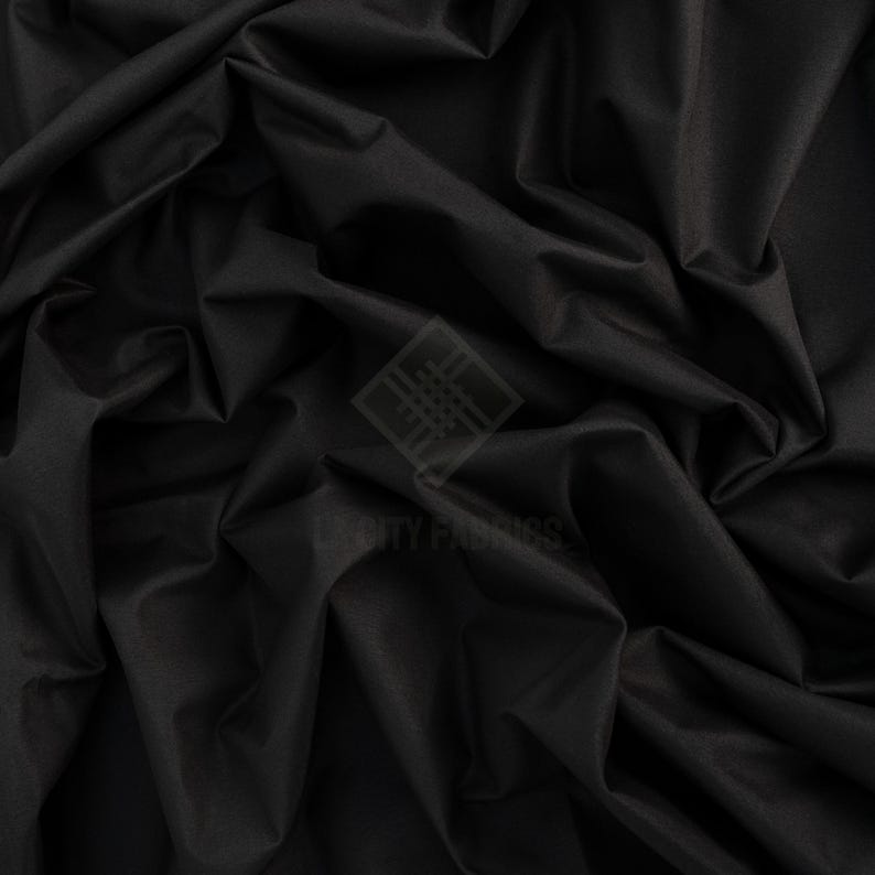 May include: A solid black fabric with a smooth, matte texture and deep, flowing folds. A semi-transparent watermark logo featuring a geometric diamond shape and the text CITY FABRICS is centered on the surface of the dark, draped material.