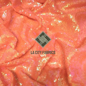 May include: A close-up shot of a coral-coloured fabric covered in sequins. The fabric is draped, creating folds and shadows. The text "LA CITY FABRICS" is visible. The sequins reflect light, adding a shimmering effect. Ideal for fashion and craft projects.