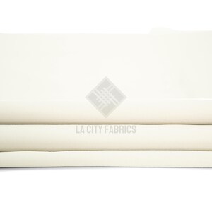 May include: A folded piece of solid off-white fabric with a smooth, matte texture. The fabric is layered in horizontal folds, creating a clean, uniform appearance. A watermark logo with the text LA CITY FABRICS is centered on the fabric.