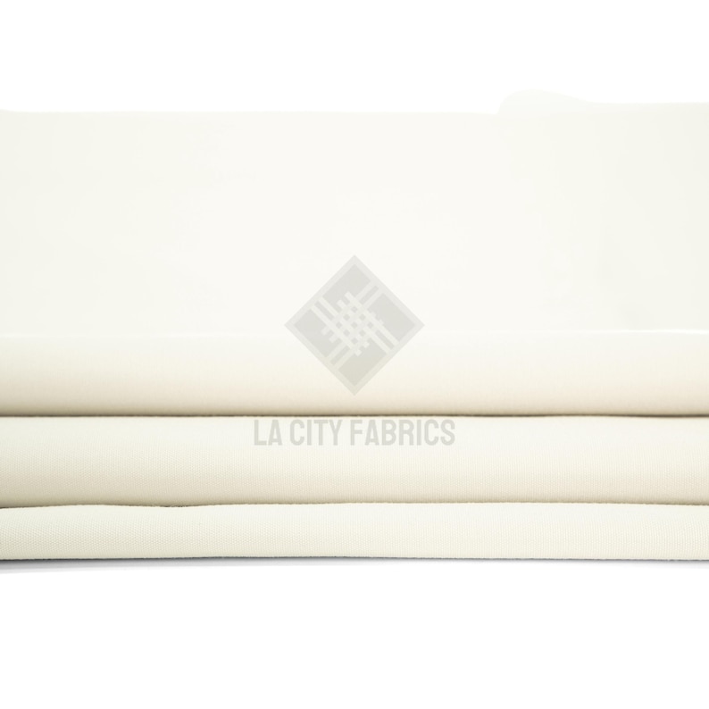May include: A folded piece of solid off-white fabric with a smooth, matte texture. The fabric is layered in horizontal folds, creating a clean, uniform appearance. A watermark logo with the text LA CITY FABRICS is centered on the fabric.