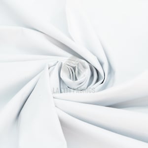 May include: A solid white fabric with a smooth, matte texture, arranged in a central spiral fold that creates soft, radiating pleats. The text LA CITY FABRICS is printed in a light gray color near the center of the fabric's spiral.