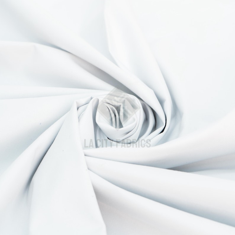 May include: A solid white fabric with a smooth, matte texture, arranged in a central spiral fold that creates soft, radiating pleats. The text LA CITY FABRICS is printed in a light gray color near the center of the fabric's spiral.