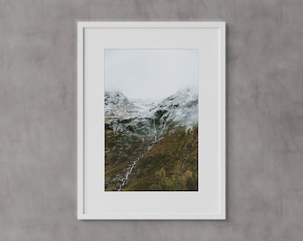 May include: A framed photograph of a mountain range with snow-capped peaks and a valley covered in green vegetation. The sky is cloudy and the overall tone of the image is muted.
