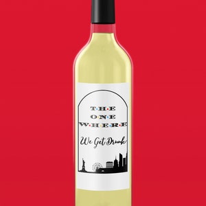 Op de afbeelding: Een fles witte wijn met een etiket waarop "The One Where We Get Drunk" staat in zwarte tekst met gekleurde stippen. Het etiket toont ook een zwart silhouet van de skyline van New York.