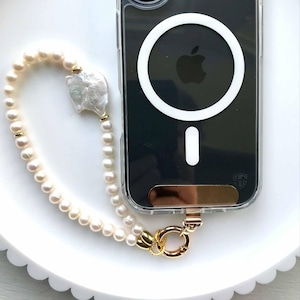 May include: Clear phone case with a pearl beaded strap. The strap features a large baroque pearl and gold-colored accents. The phone case has a gold-colored kickstand and a white MagSafe ring.