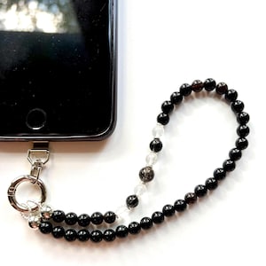 Könnte beinhalten: Ein schwarzes Smartphone mit einer Perlen-Handschlaufe. Die Schlaufe besteht aus einem wiederkehrenden Muster kleiner, polierter schwarzer Perlen, die mit einigen klaren, durchsichtigen Perlen und einem silberfarbenen Karabinerhaken versehen sind.