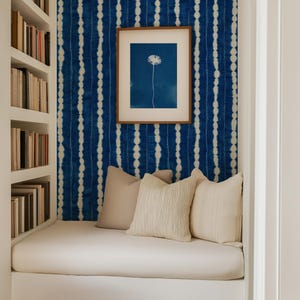 May include: A cozy reading nook featuring a built-in bench with three beige throw pillows. The wall is covered in blue and white tie-dye patterned wallpaper. A framed print of a white dandelion on a blue background hangs centered on the wall.