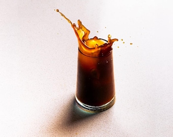 Peut inclure: Un verre cylindrique transparent rempli de café glacé brun foncé, avec une éclaboussure de liquide dynamique jaillissant du sommet. La boisson est d'une couleur brun profond et le verre présente une base épaisse et transparente.