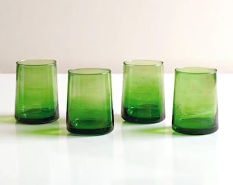 Peut inclure: Quatre verres à boire teintés en vert. Les verres ont une forme cylindrique avec un design légèrement conique. La couleur passe d'un vert plus foncé à la base à une teinte plus claire vers le haut. Ils sont disposés sur une surface blanche.