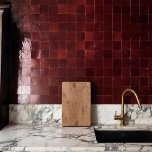 May include: A kitchen interior featuring a marble countertop with a black sink and a gold faucet. A wooden cutting board rests on the countertop. The wall is covered in square, dark red tiles. A wooden cabinet is on the left.