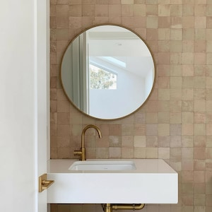 Puede incluir: Un tocador de baño moderno con un espejo redondo con marco dorado, un lavabo blanco y un grifo dorado. La pared está alicatada con baldosas cuadradas de tonos tierra. Una puerta blanca y un tirador dorado son visibles a la izquierda.